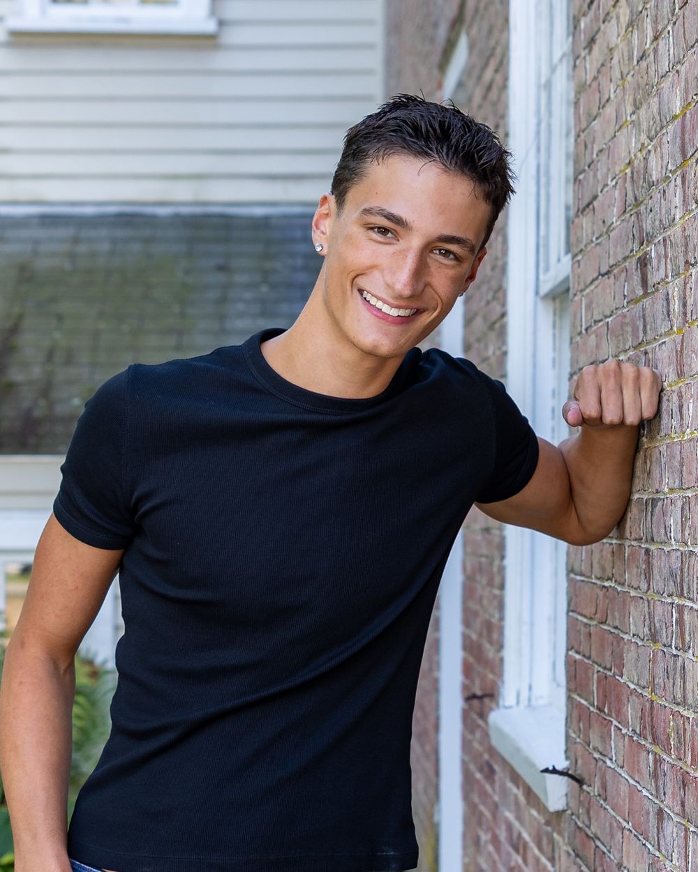 A high school senior in a black t-shirt posing casually against a brick wall outdoors during yearbook portraits in Sudbury, MA.