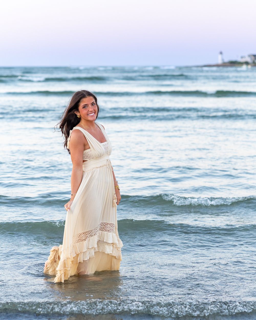 High school senior wearing a flowing white dress stands in the ocean waves with a lighthouse visible in the background during yearbook photo