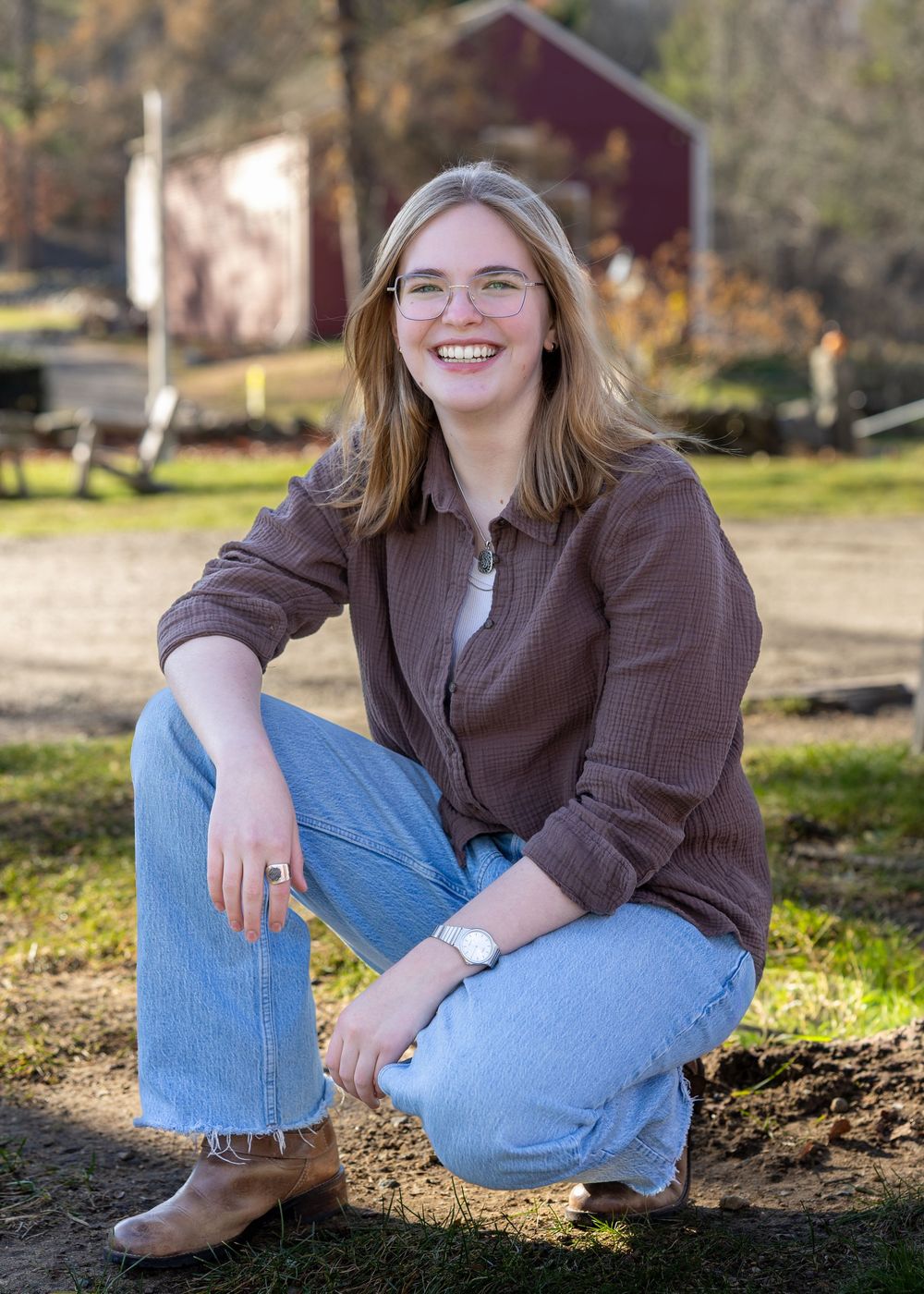 High school senior poses in a brown shirt and jeans sitting on the ground in natural lighting by a red barn at Wayside Inn in Sudbury, MA.