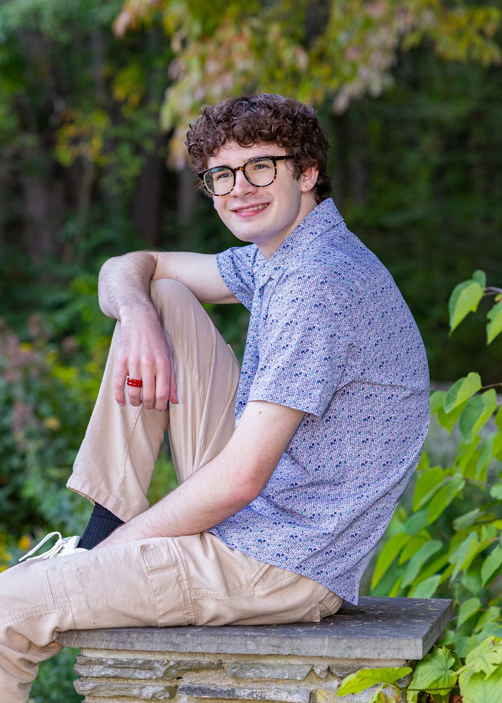 High school senior in a blue floral shirt & khaki pants sits on a stone ledge in a garden setting in a yearbook photo session in Sudbury, MA