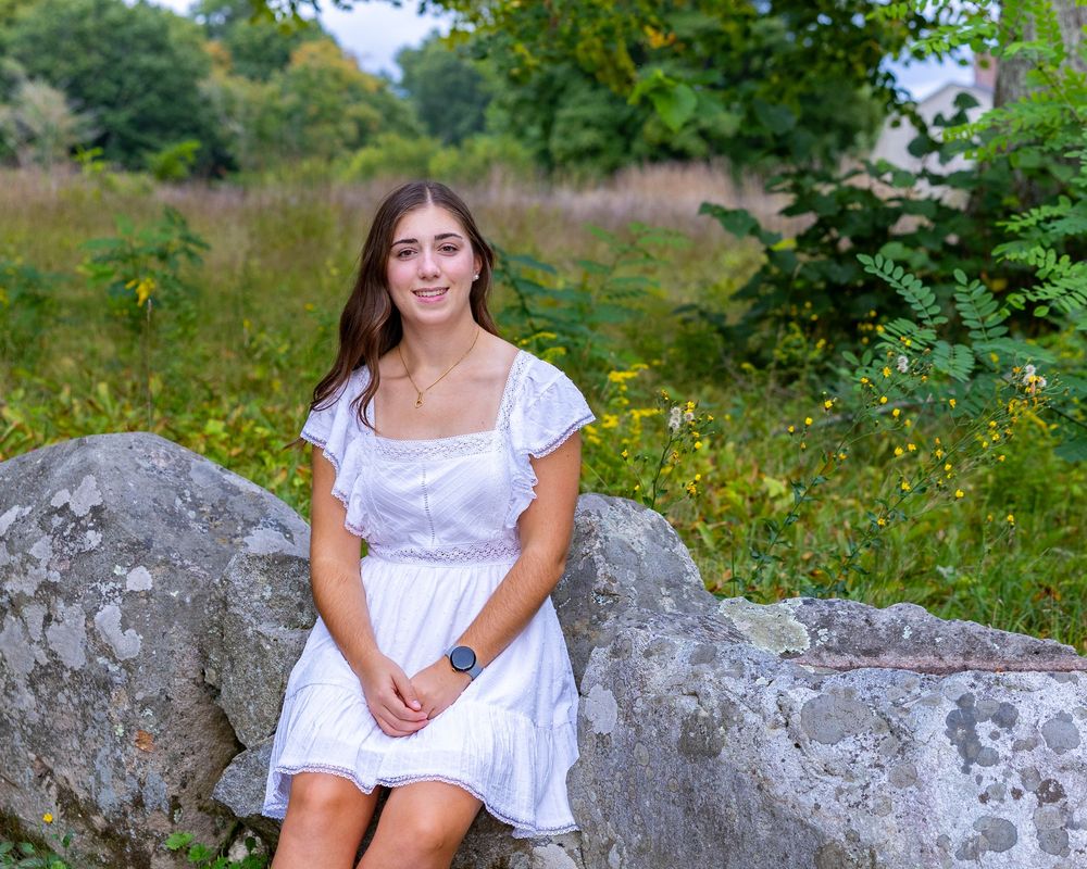 High school senior in white dress sits on a large gray rock surrounded by wildflowers and greenery in a summer meadow during yearbook photos
