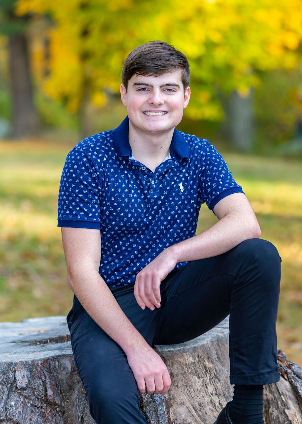 High school senior in a navy polka dot shirt sits casually on a tree stump in an autumn setting with golden foliage at Grist Mill in Sudbury