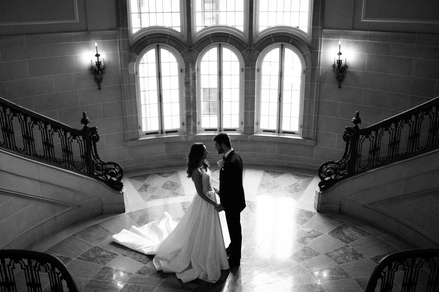 A dramatic black and white photograph of an elegant staircase in a historic building with arched windows and natural light.