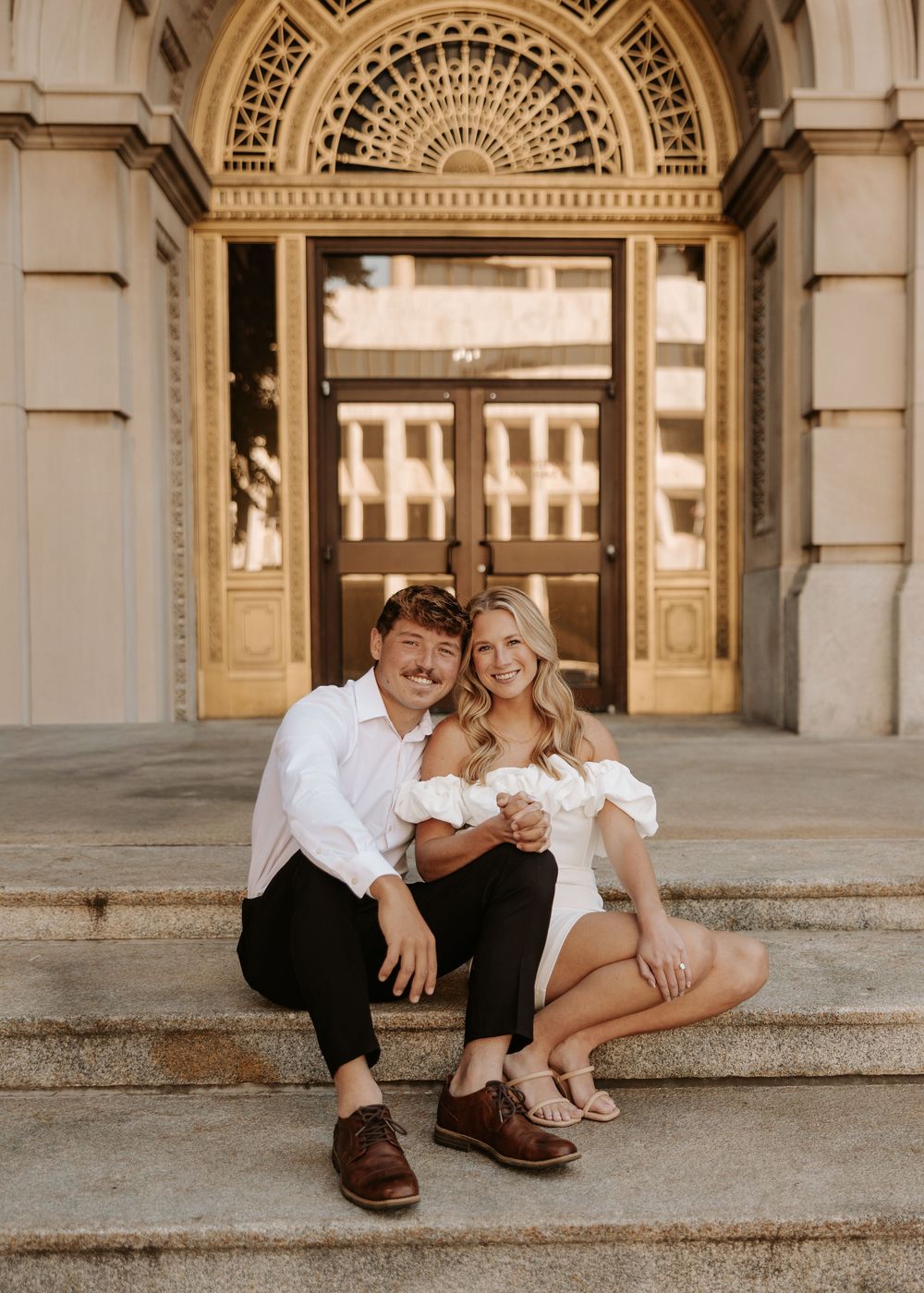 A couple sits on ornate stone steps in front of grand architectural doorway with decorative fanlight above.