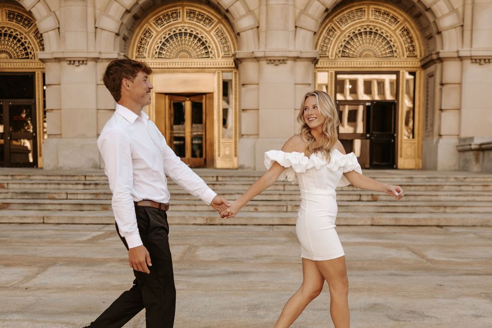 A couple dances joyfully on stone steps outside an ornate building with arched doorways and classical architecture.