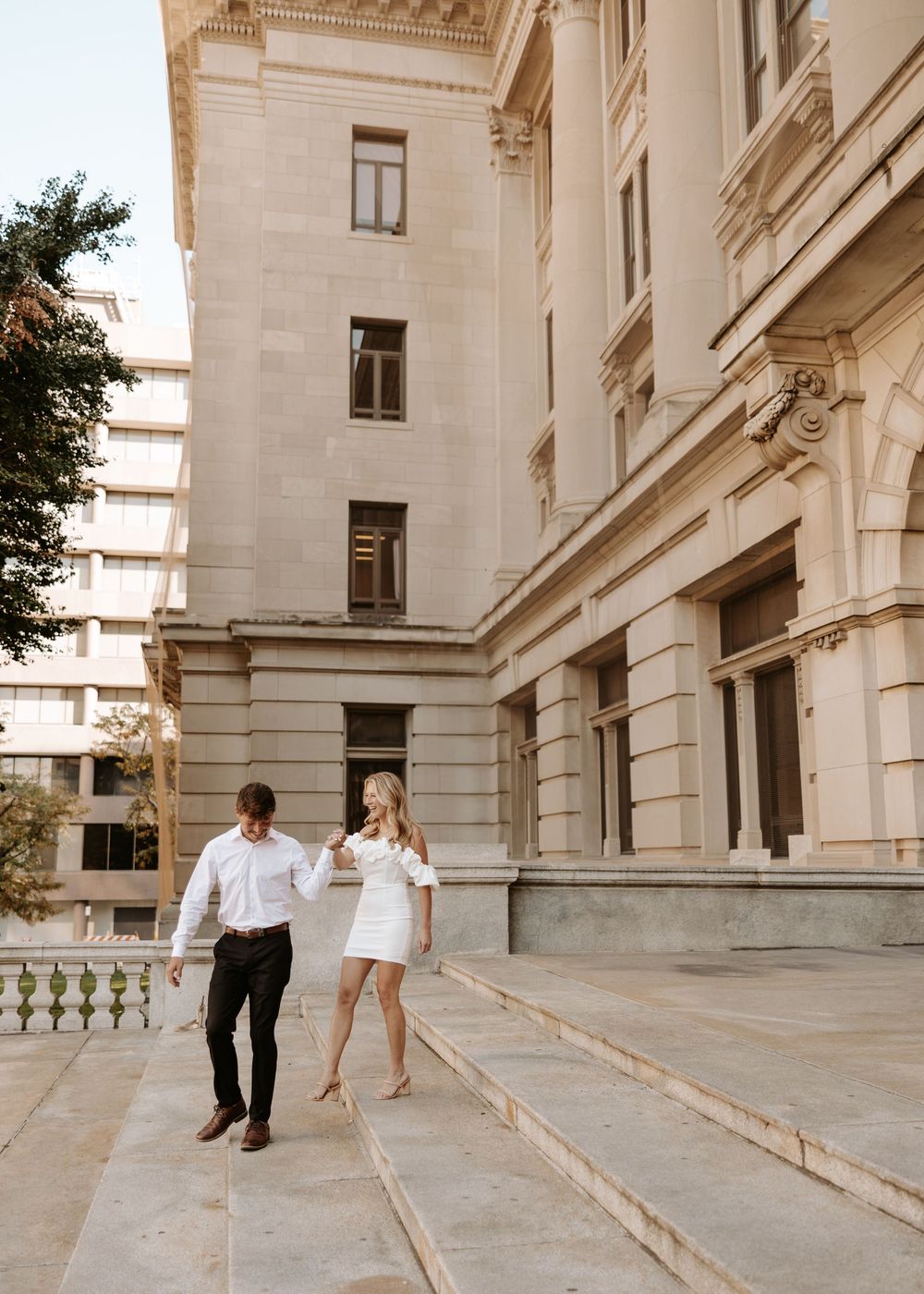A couple in white outfits playfully walks up stone steps along a grand historic building exterior.