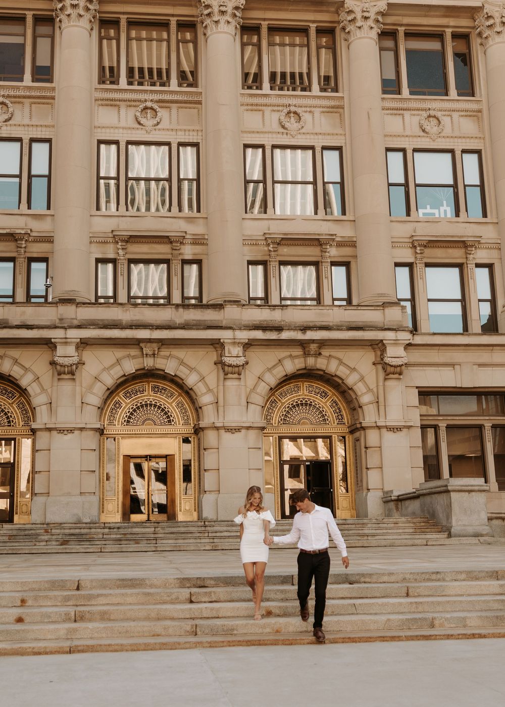 A couple walks up grand stone steps towards ornate arched entrances of a historic building.