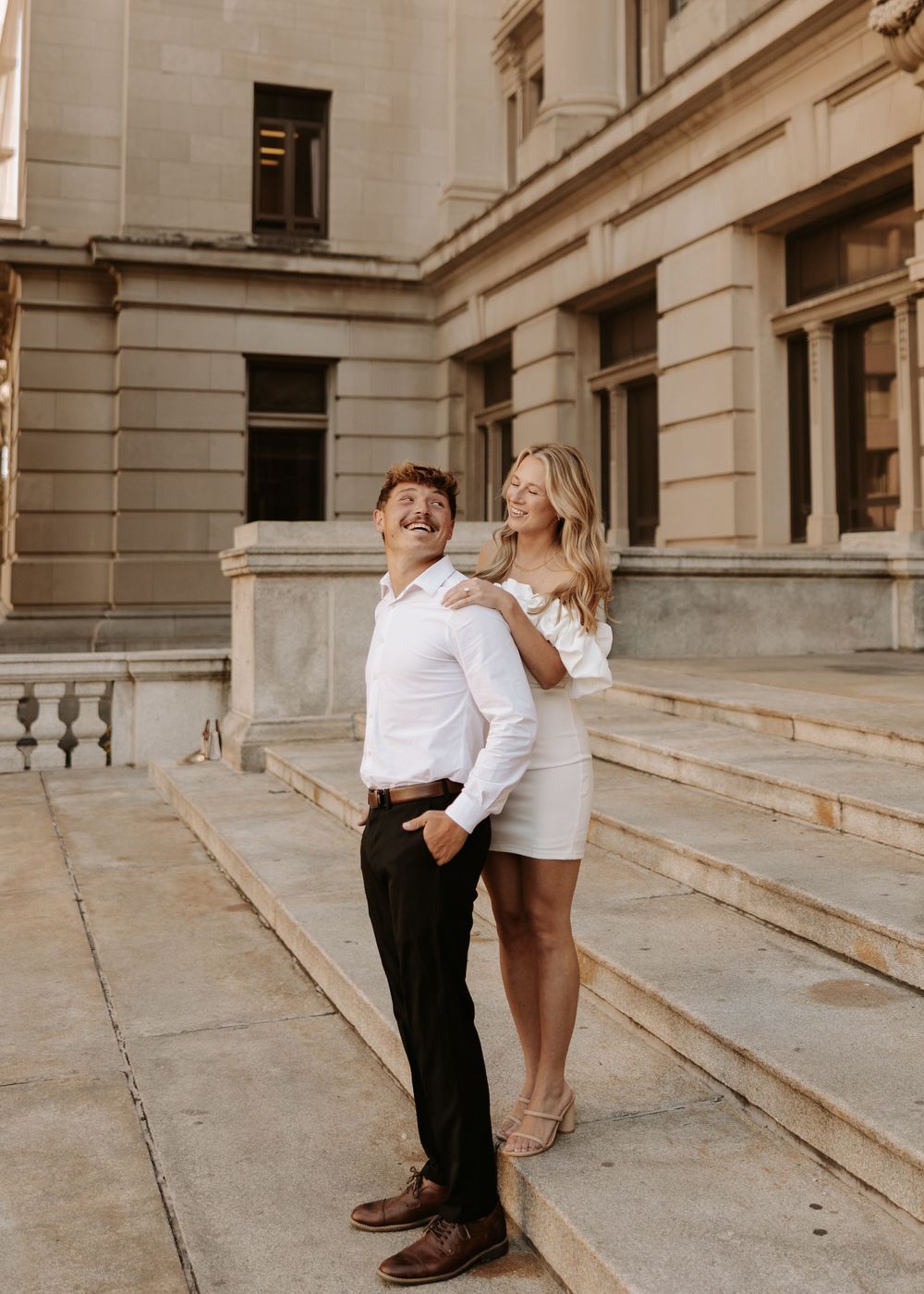 A romantic couple in matching white outfits dances together on elegant stone steps by a classical building.