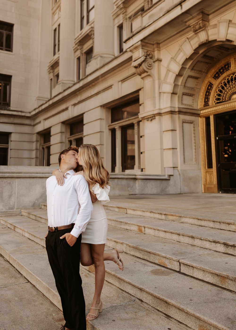 A couple in formal attire embraces on elegant stone steps outside a historic building.