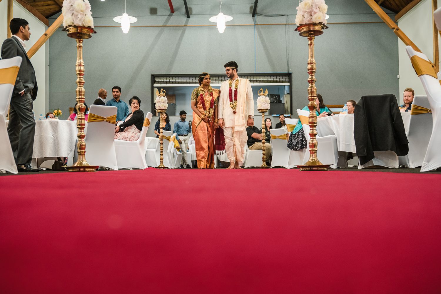 Traditional Indian wedding ceremony on a red carpet stage with decorative lamps and guests seated in the background.
