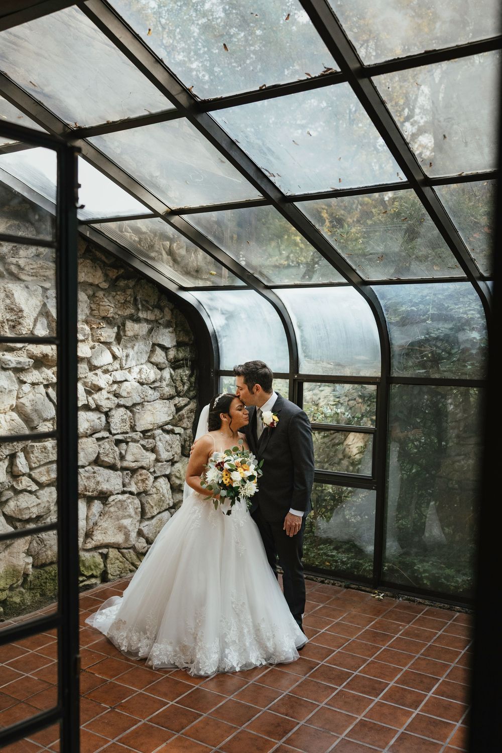 A couple shares an intimate moment in a rustic greenhouse with stone walls and terracotta floors.