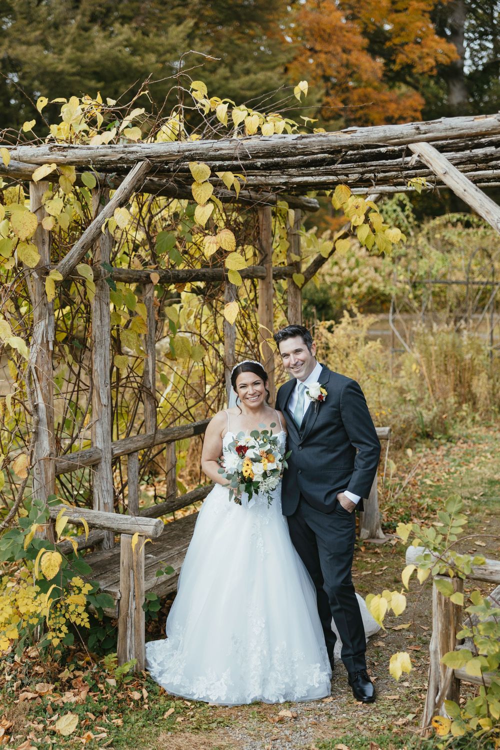 A rustic autumn wedding photo session under a wooden arbor with yellow leaves and fall foliage in the background.