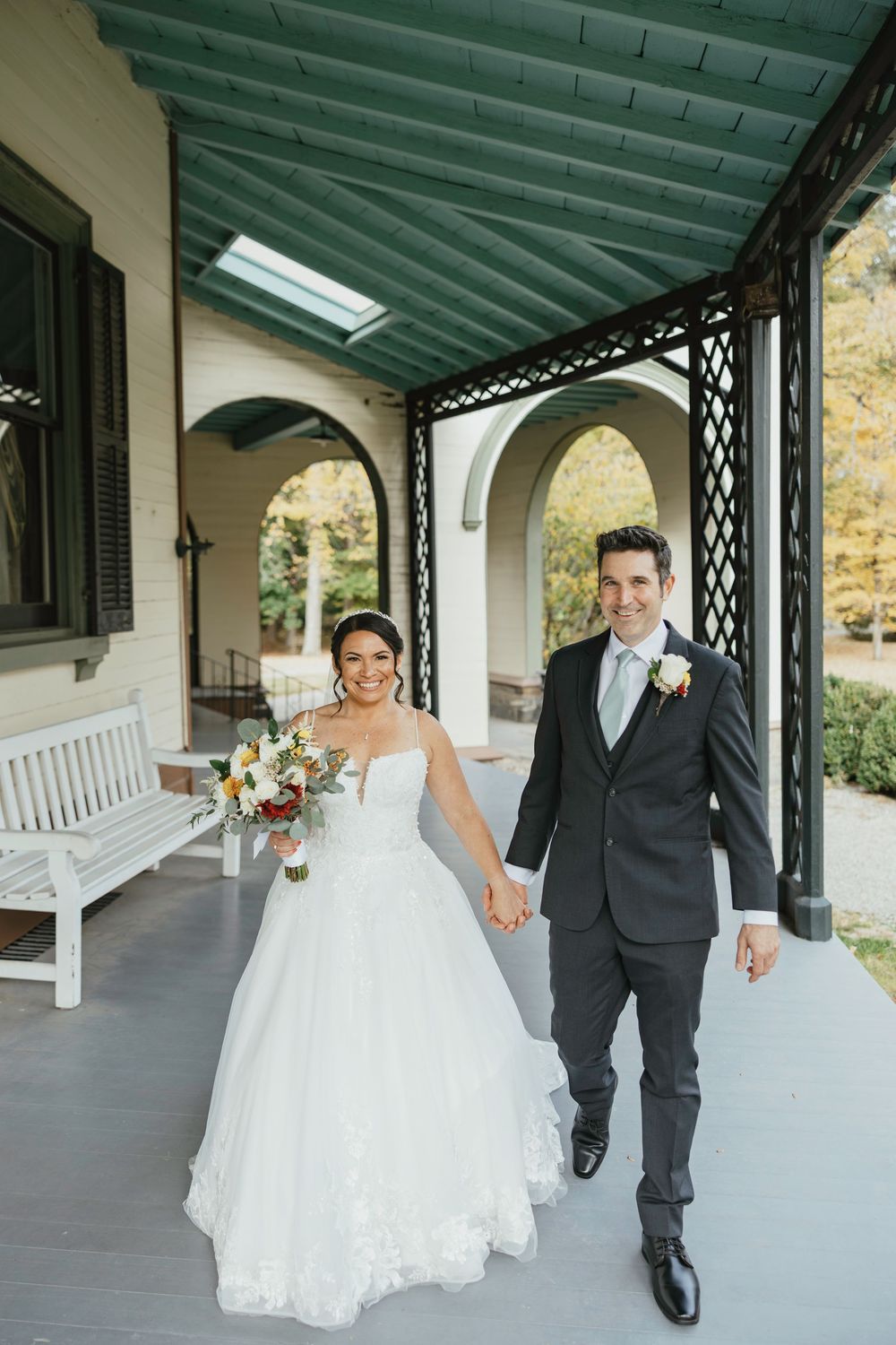 Couple walking together on a covered porch with white columns and stone walls during their wedding celebration.