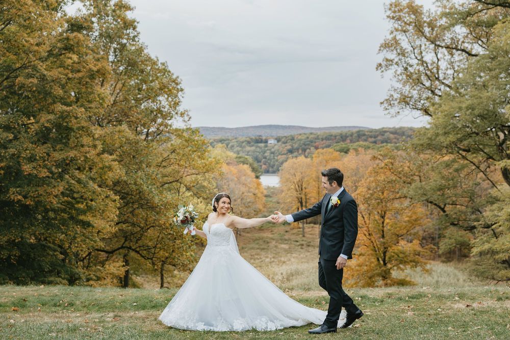 A couple in wedding attire poses together in a scenic autumn landscape with golden foliage and rolling hills.