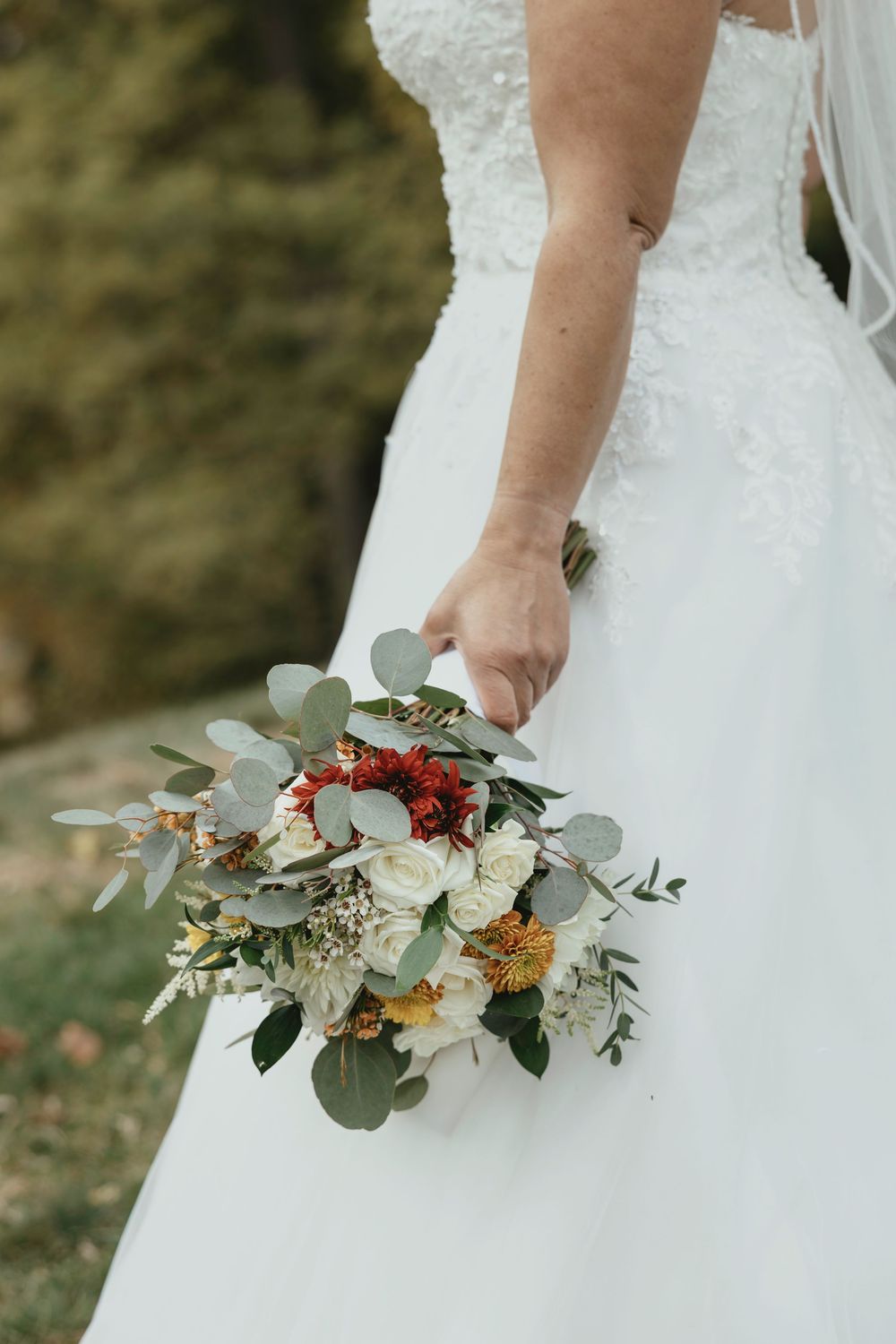 A bride holds a rustic autumn wedding bouquet with eucalyptus, white flowers and orange accents against her white dress.