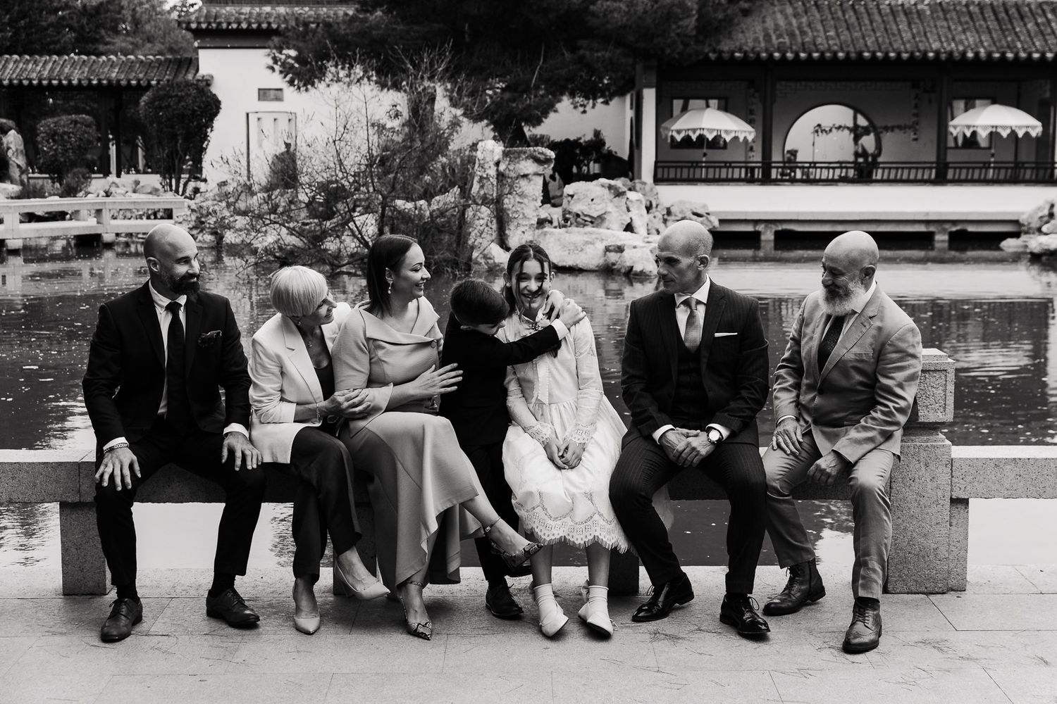 A group of people sit casually in a row on a ledge or bench in a black and white photograph.