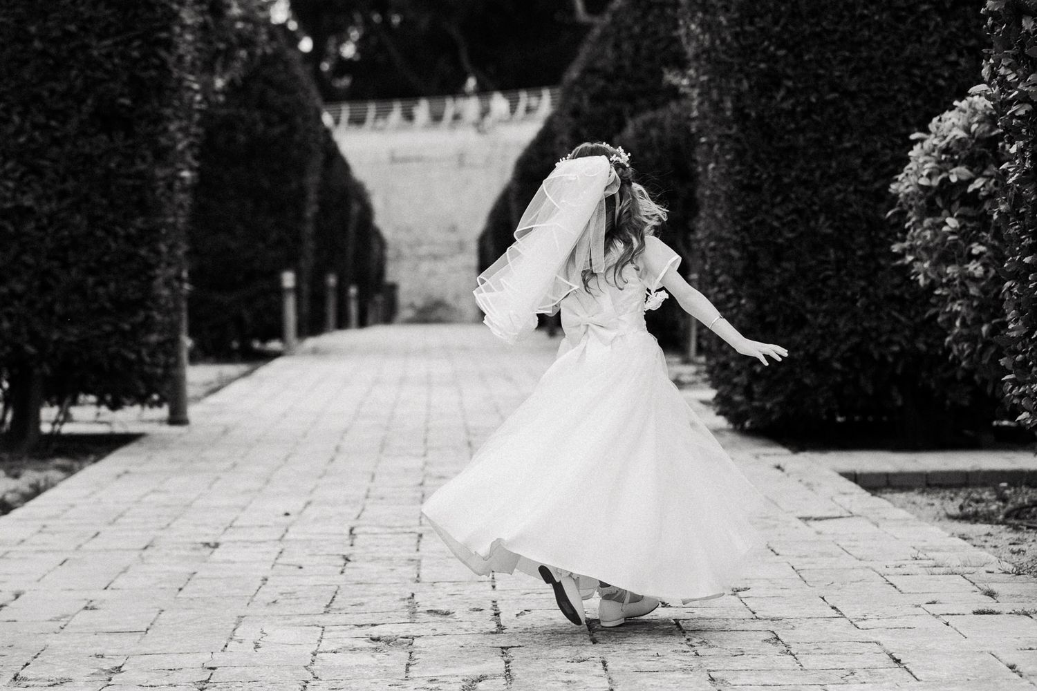 A bride in a flowing white wedding dress twirls gracefully on a stone pathway surrounded by manicured gardens.