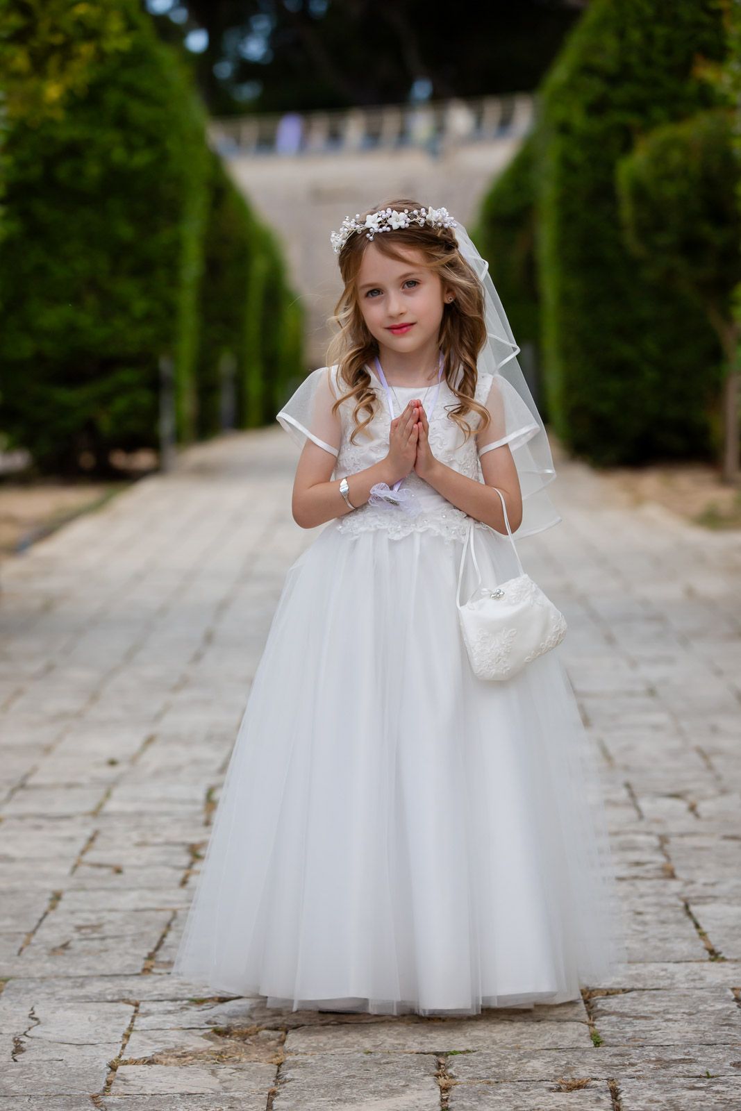 Young child in white First Communion dress with tiara and purse standing on stone path between manicured hedges.