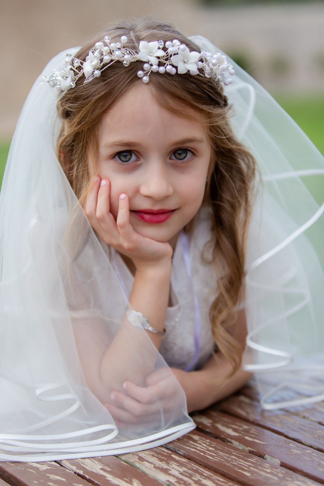Child wearing delicate white veil and floral crown rests chin on hand in communion portrait.