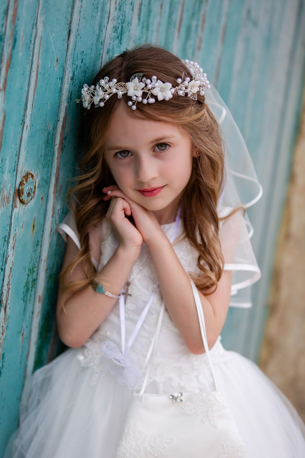 Young girl in white dress and floral headpiece poses against turquoise door for first communion portrait.