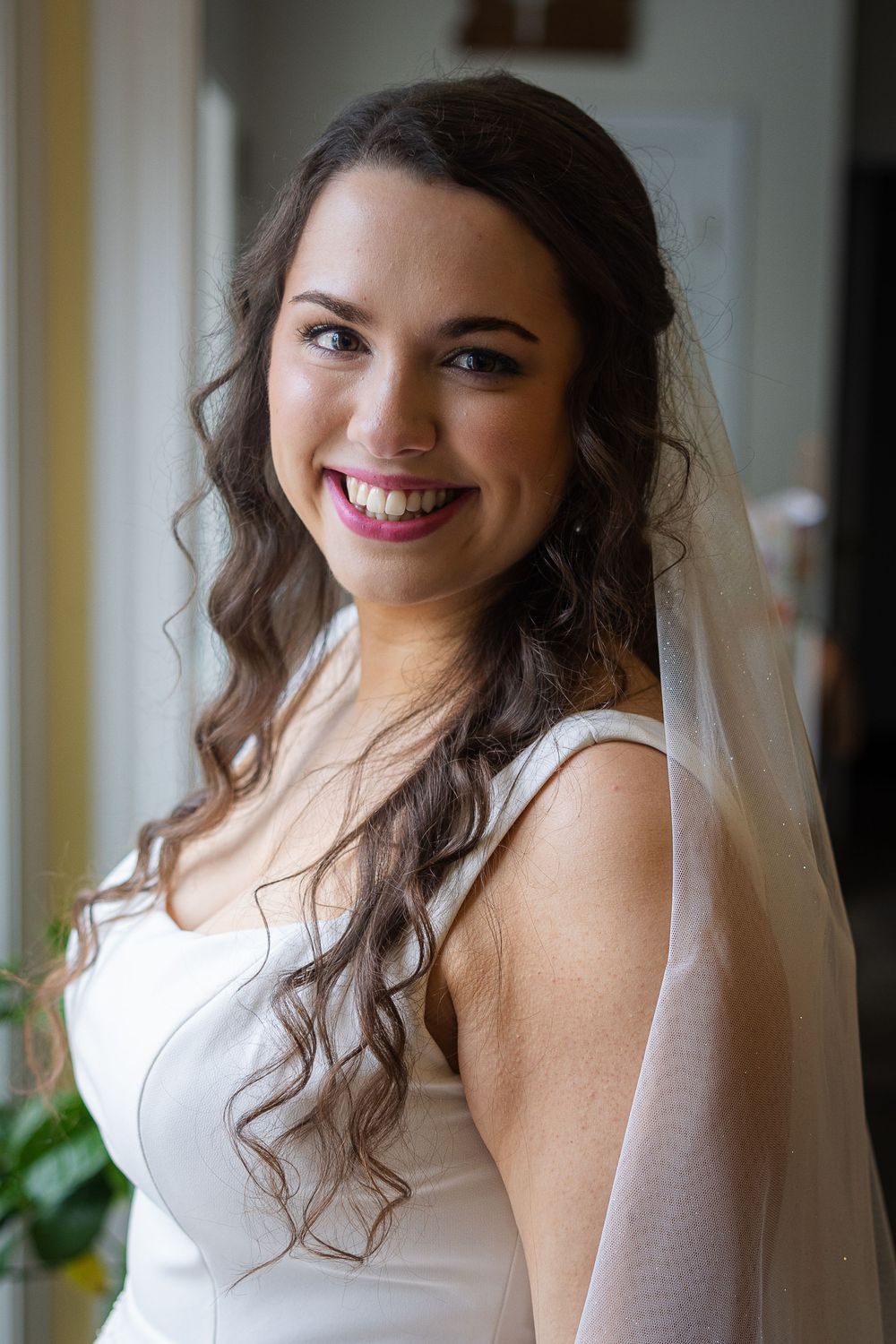 A bride in a white wedding dress poses with curled hair and a veil in an elegant indoor setting.
