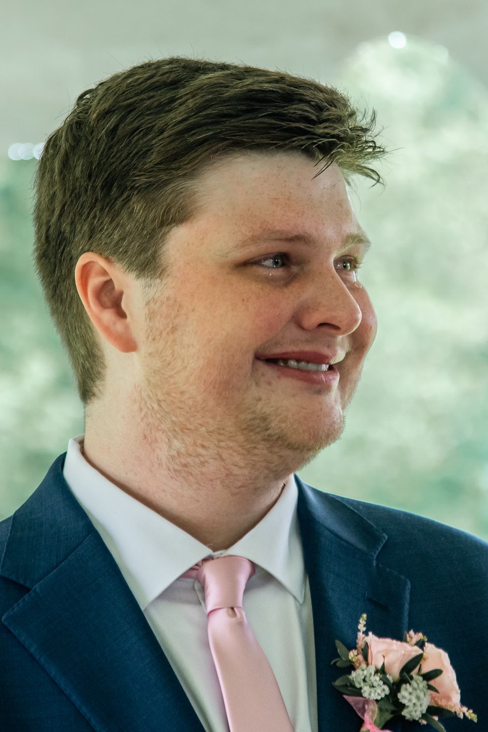 A groomsman in a navy suit and pink tie smiles while wearing a pink and white floral boutonniere.