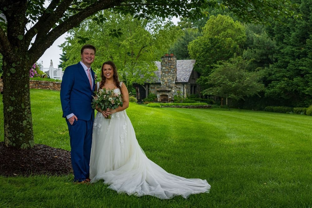A couple in wedding attire stands together on a lush green lawn surrounded by trees on their special day.