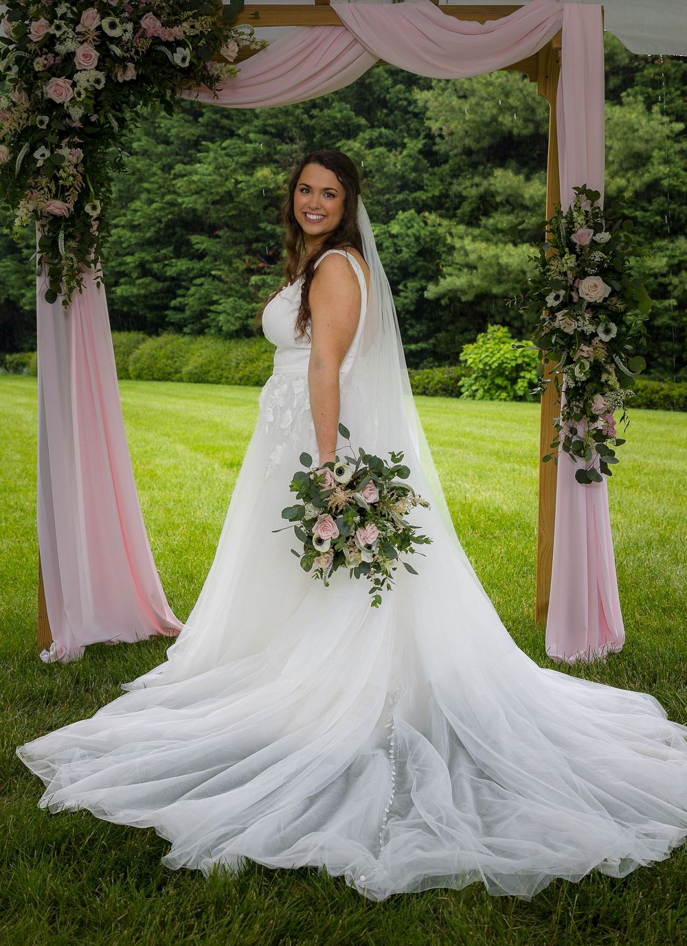 A bride in a flowing white wedding gown poses elegantly in a garden setting with pink drapery and floral decorations.