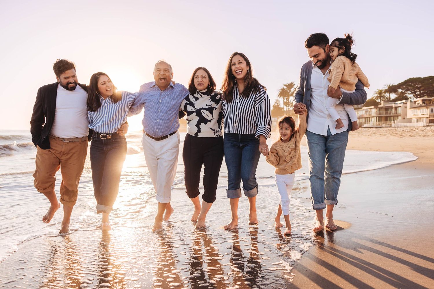 Family walking together on beach at sunset with waves splashing around their feet as golden light shines through.
