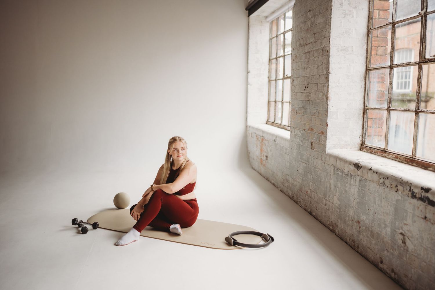 Woman in red activewear sitting on yoga mat with pilates ring and foam roller in bright studio.