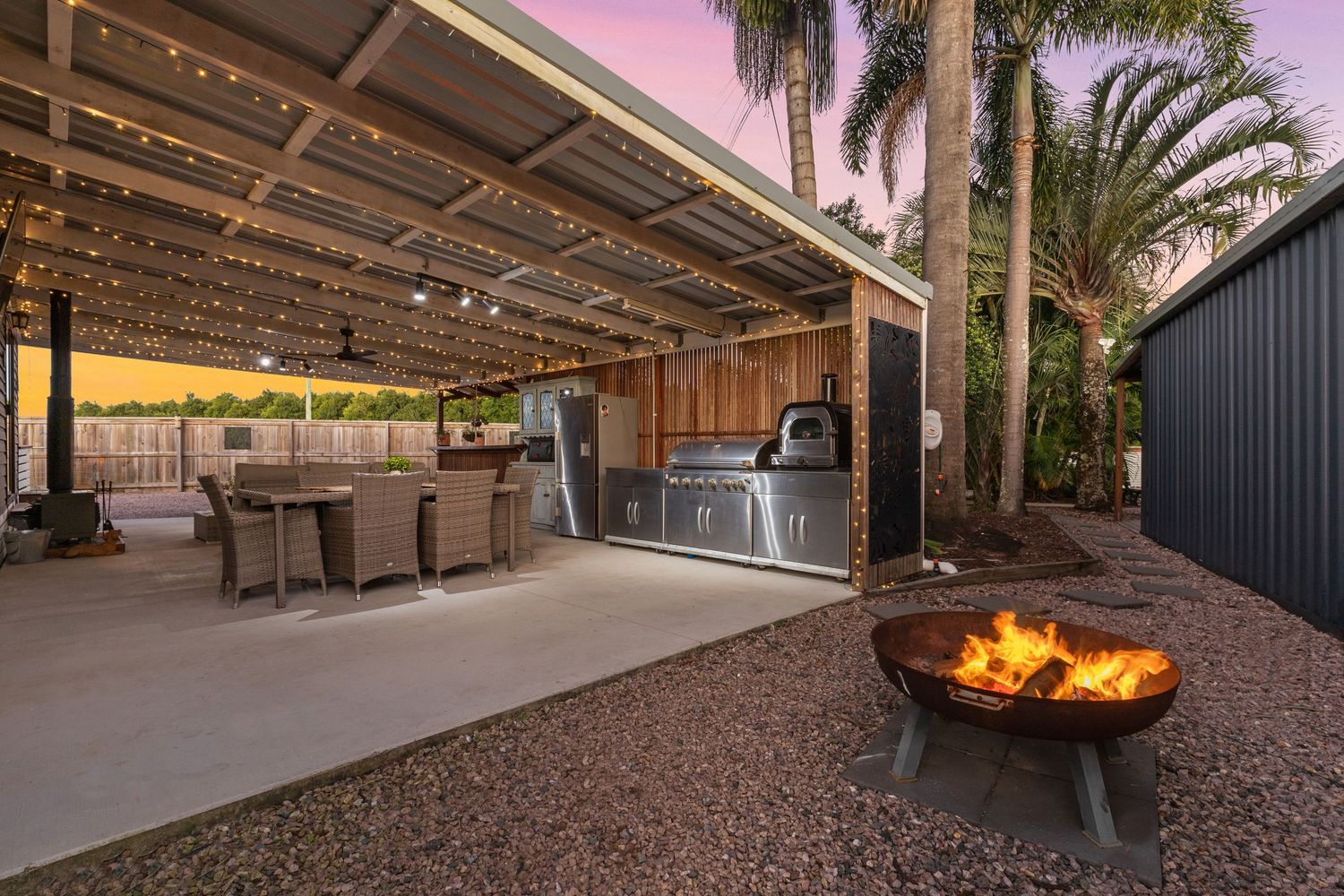 Outdoor entertaining area with pergola, BBQ kitchen, fire pit, rattan dining set, and palm trees at sunset.