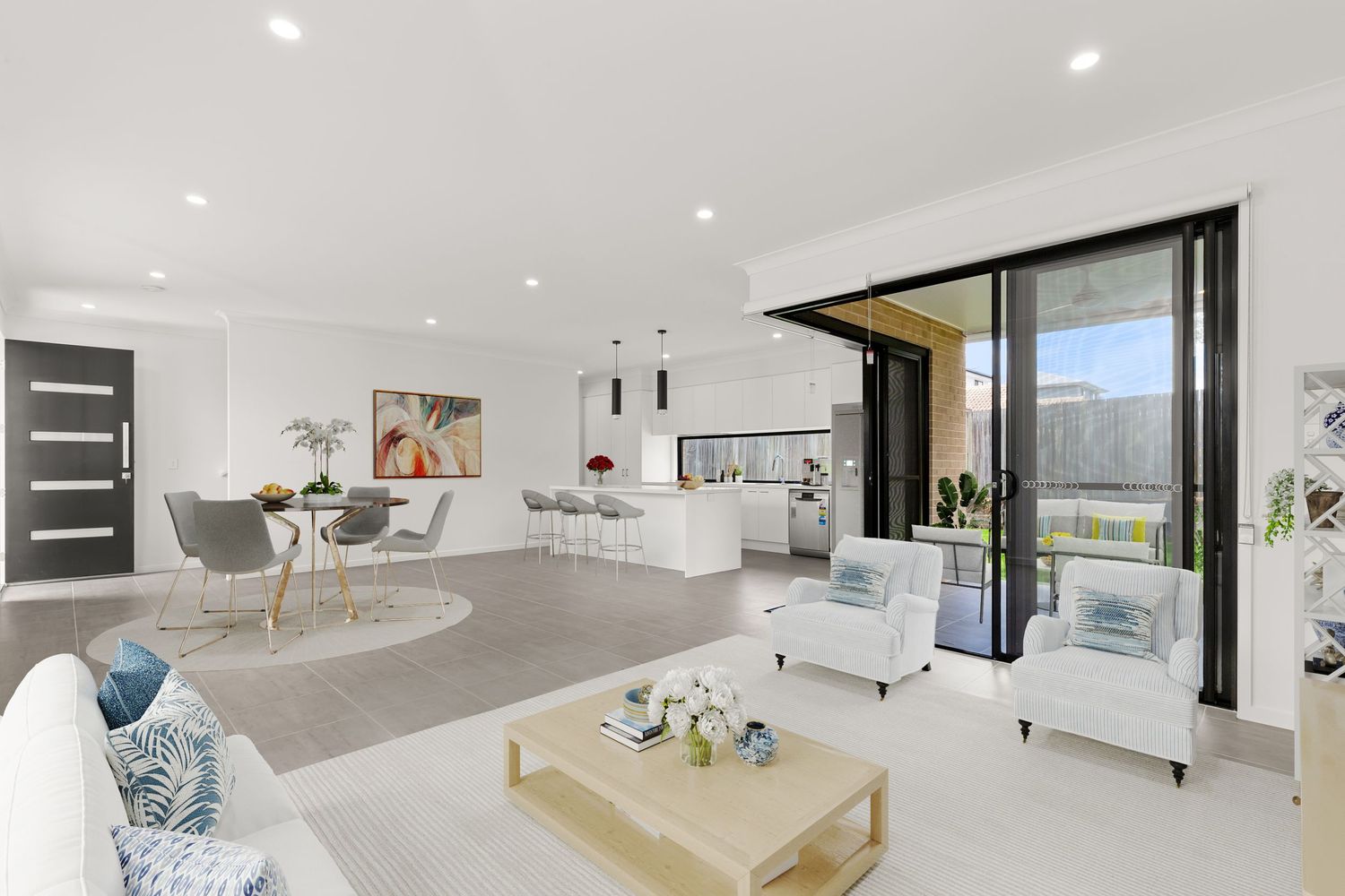 Bright open-plan living and dining area with white interiors, sliding glass doors, and modern kitchen in background.