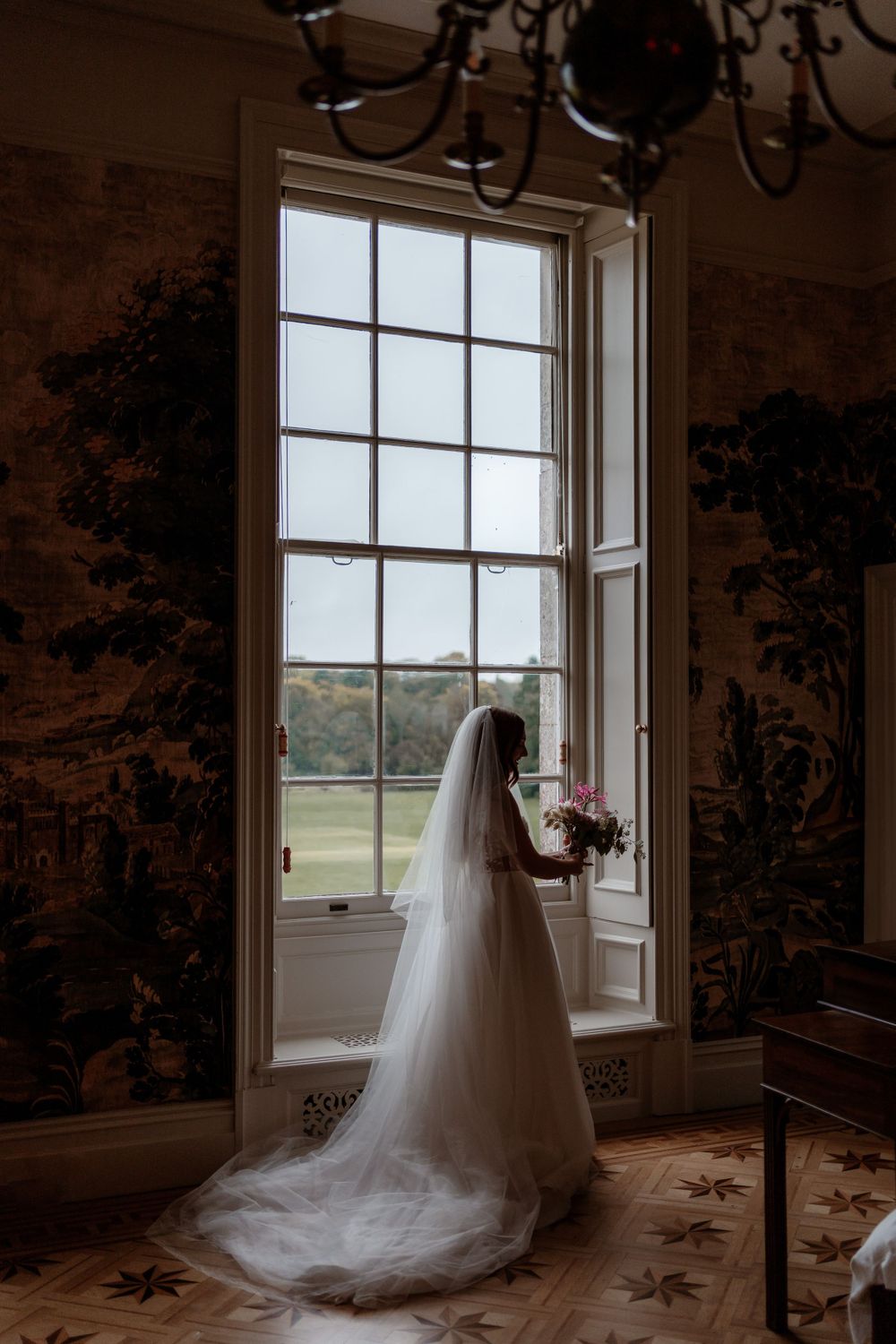 Silhouette of a wedding dress against large windows in an elegant room with wallpaper.
