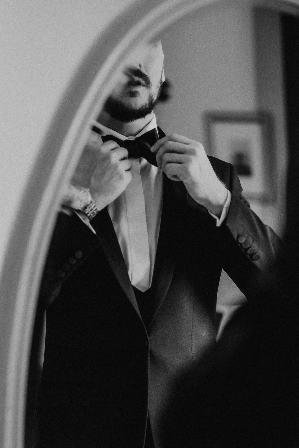 A groom adjusts his bowtie in a series of black and white mirror reflections.