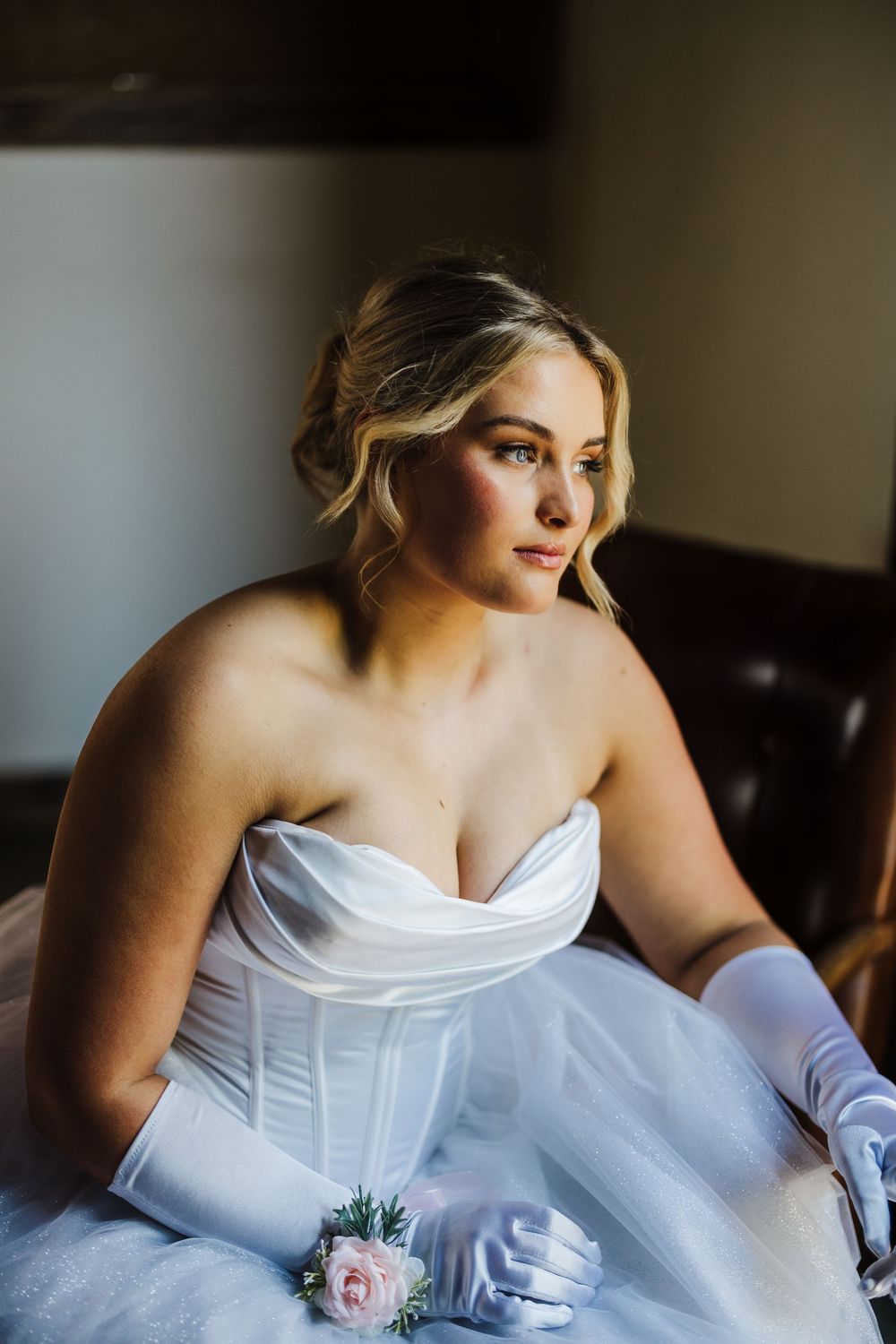 A bride in a white strapless gown with a delicate pink rose corsage poses in soft natural light.
