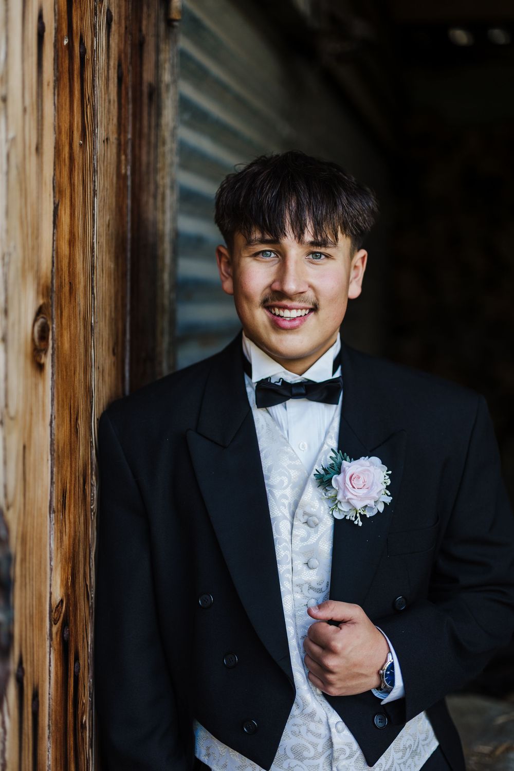 A groom in a black tuxedo with a white vest and pink boutonniere smiles while standing between wooden posts.