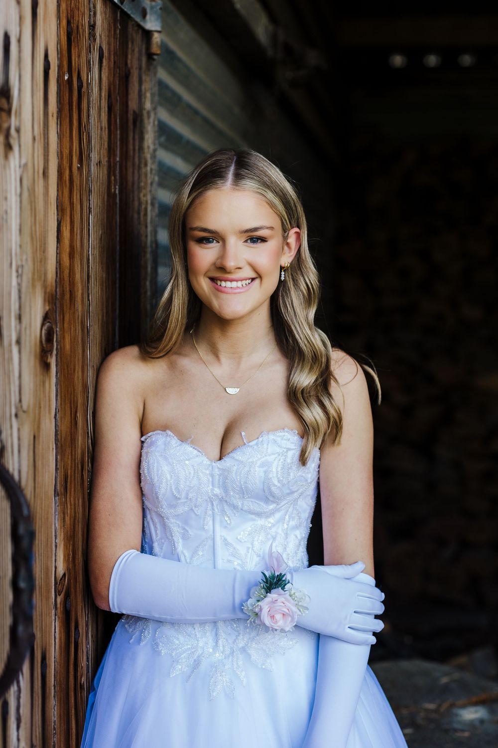 A bride in a white strapless gown and elbow-length gloves poses against a rustic wooden backdrop.