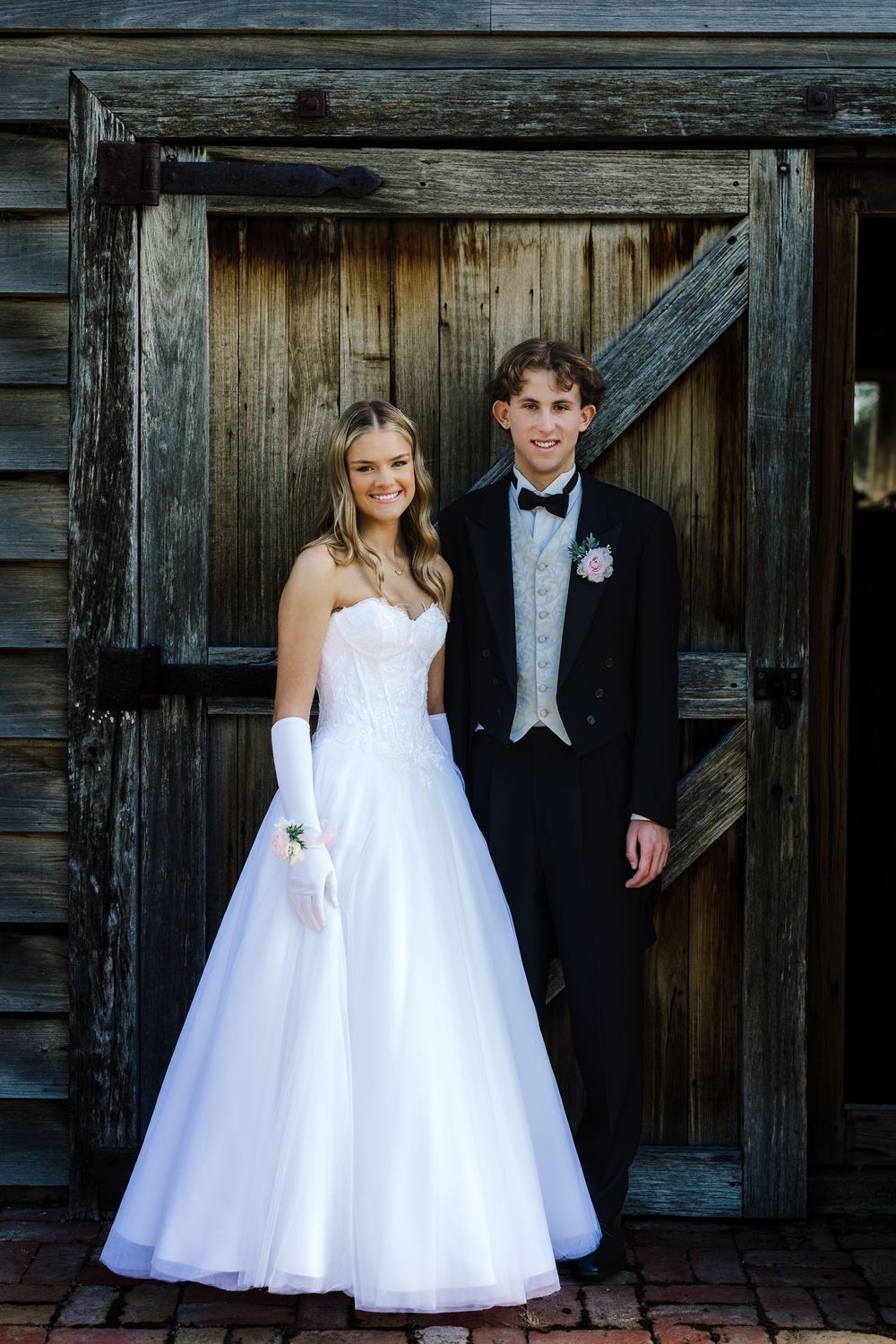 Wedding party poses in front of rustic wooden barn wearing formal white dresses and tuxedo.