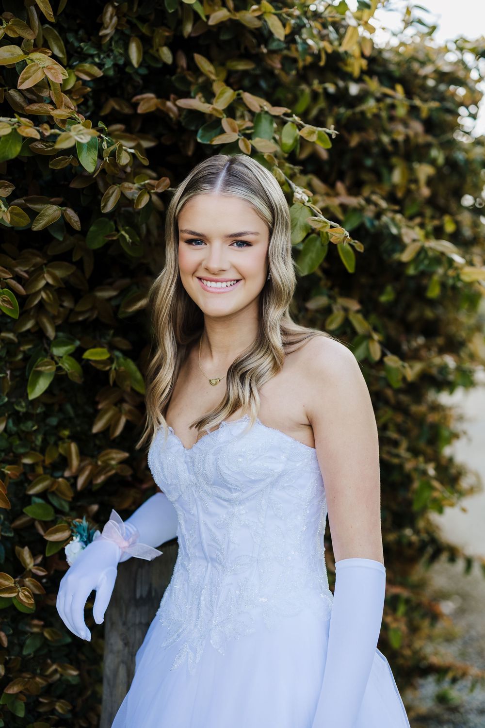 A bride in a white strapless gown and long gloves poses against a backdrop of green foliage.