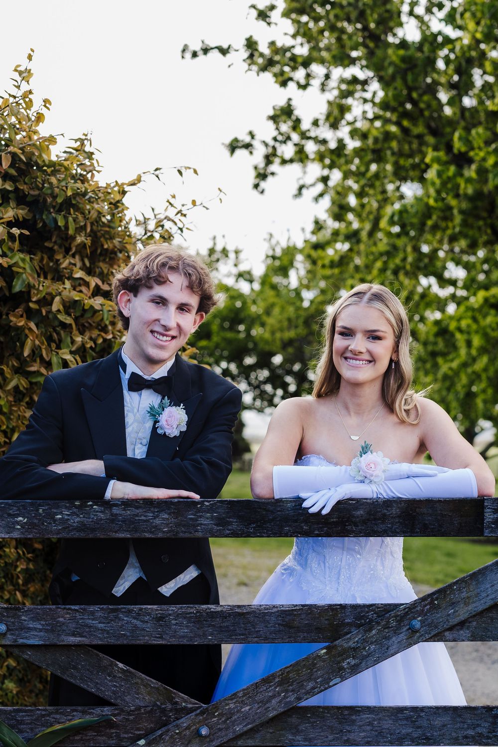 Couple in formal attire leaning on rustic wooden fence with green foliage in background.
