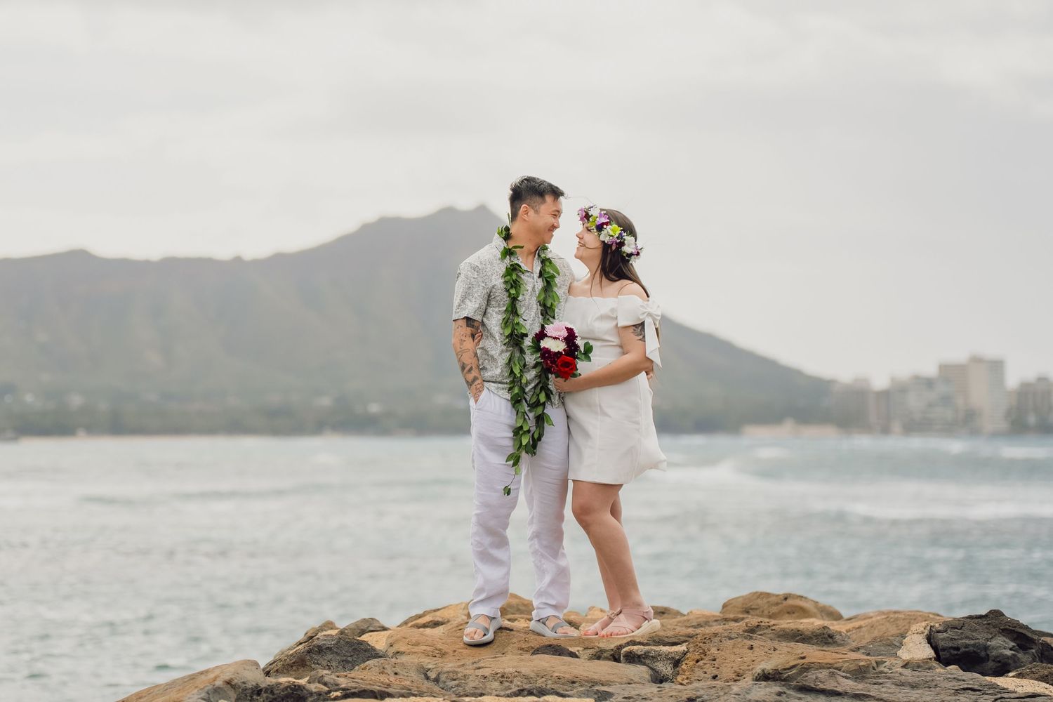 A couple in white attire poses together on rocky coastal cliffs overlooking the ocean during a photo session.