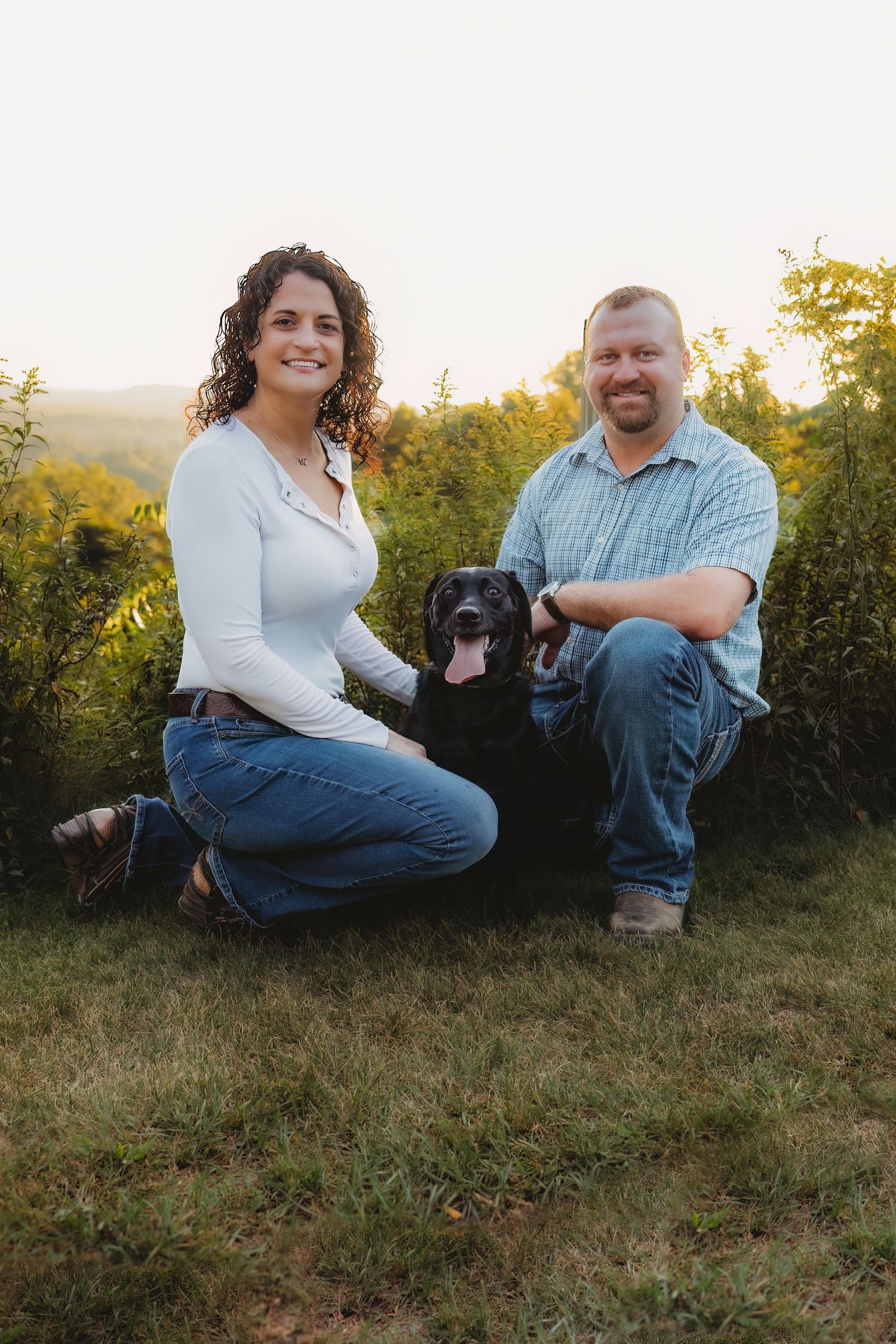 A couple sits on a grassy hillside with their black dog during golden hour sunset.