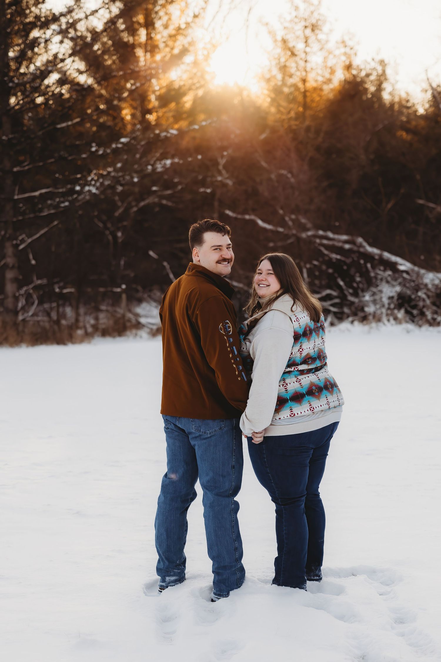 A couple stands together in a snowy winter landscape at sunset with golden light filtering through trees in the background.