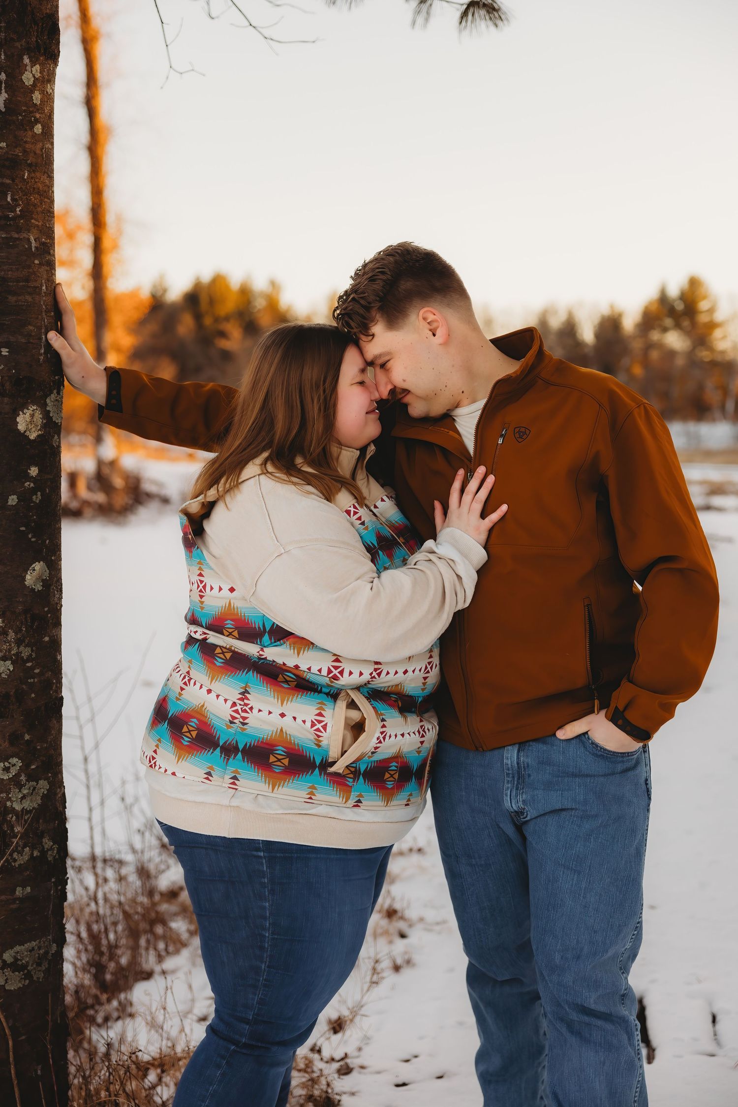 A couple shares an intimate winter moment in the snow, wearing cozy sweaters while leaning against a tree at sunset.