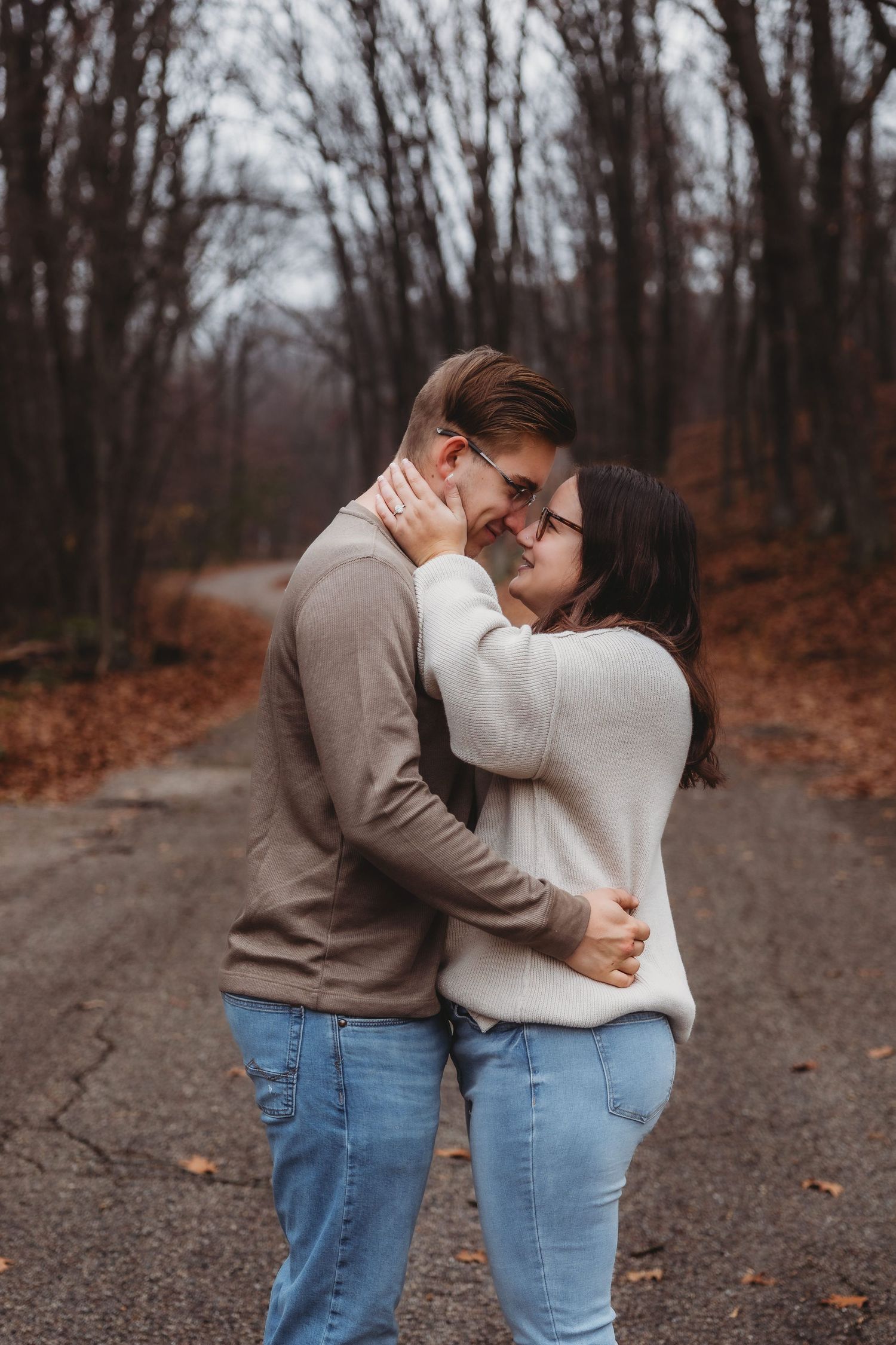 A couple embraces tenderly on a path surrounded by autumn trees with fallen leaves in the background.