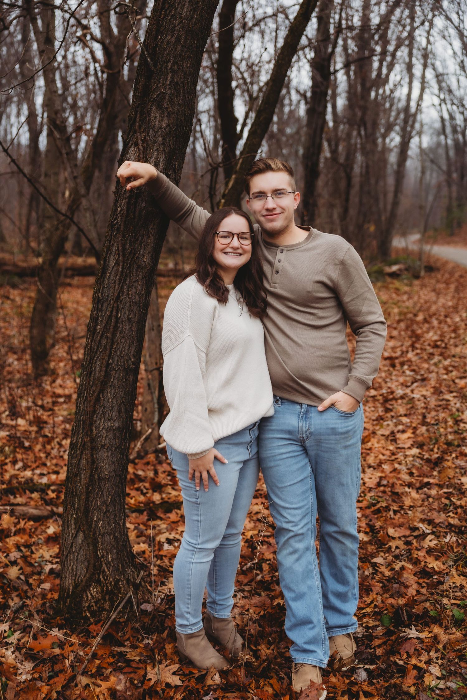 A couple poses together in a wooded area with fallen autumn leaves covering the ground.