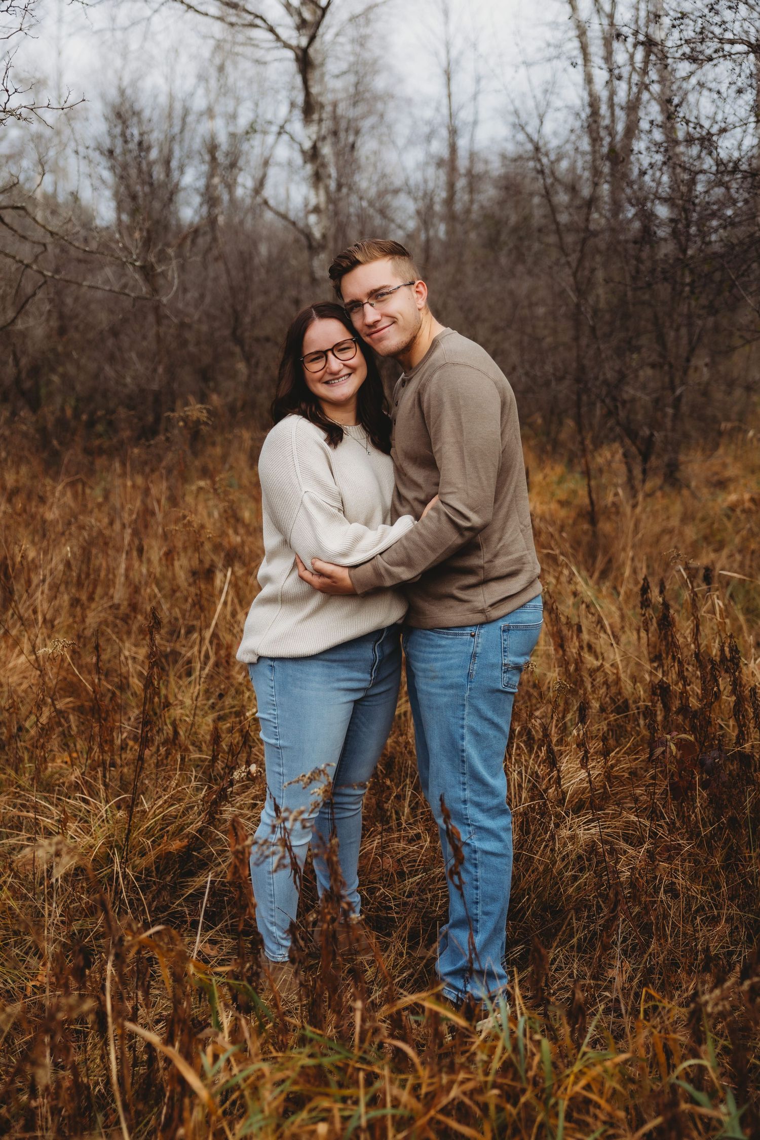 A couple in matching cream sweaters and jeans embraces while standing in tall autumn grass during a photo session.