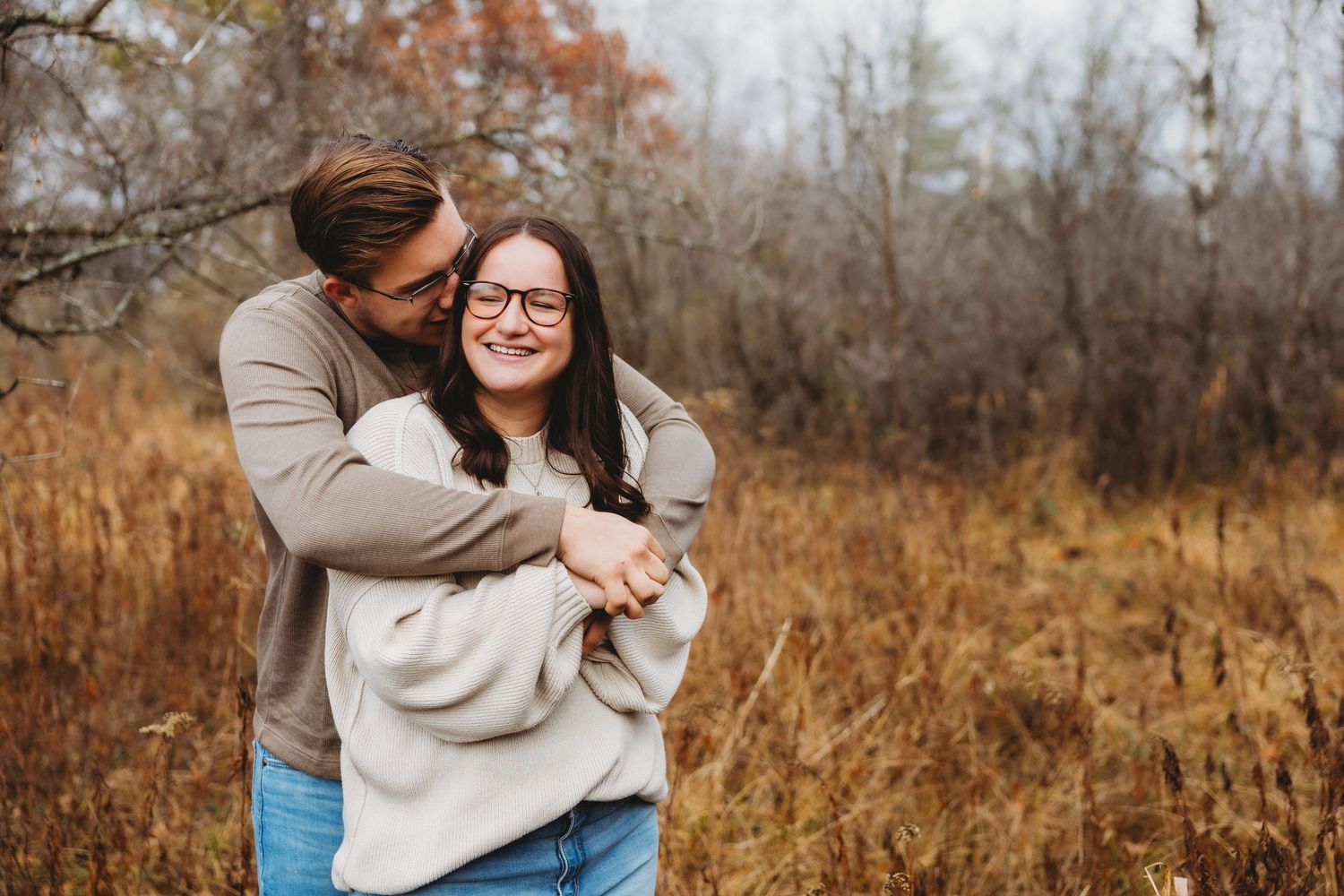 A couple embraces in a golden autumn field with tall grass and trees in the background during sunset.