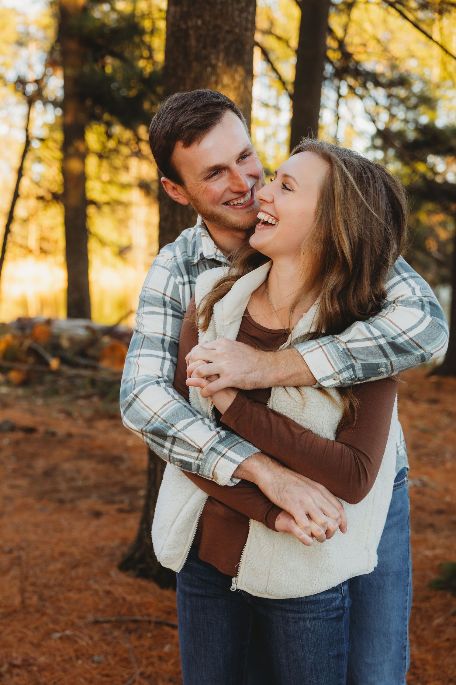 A couple embraces and laughs together during a romantic autumn photoshoot in a sunlit forest.