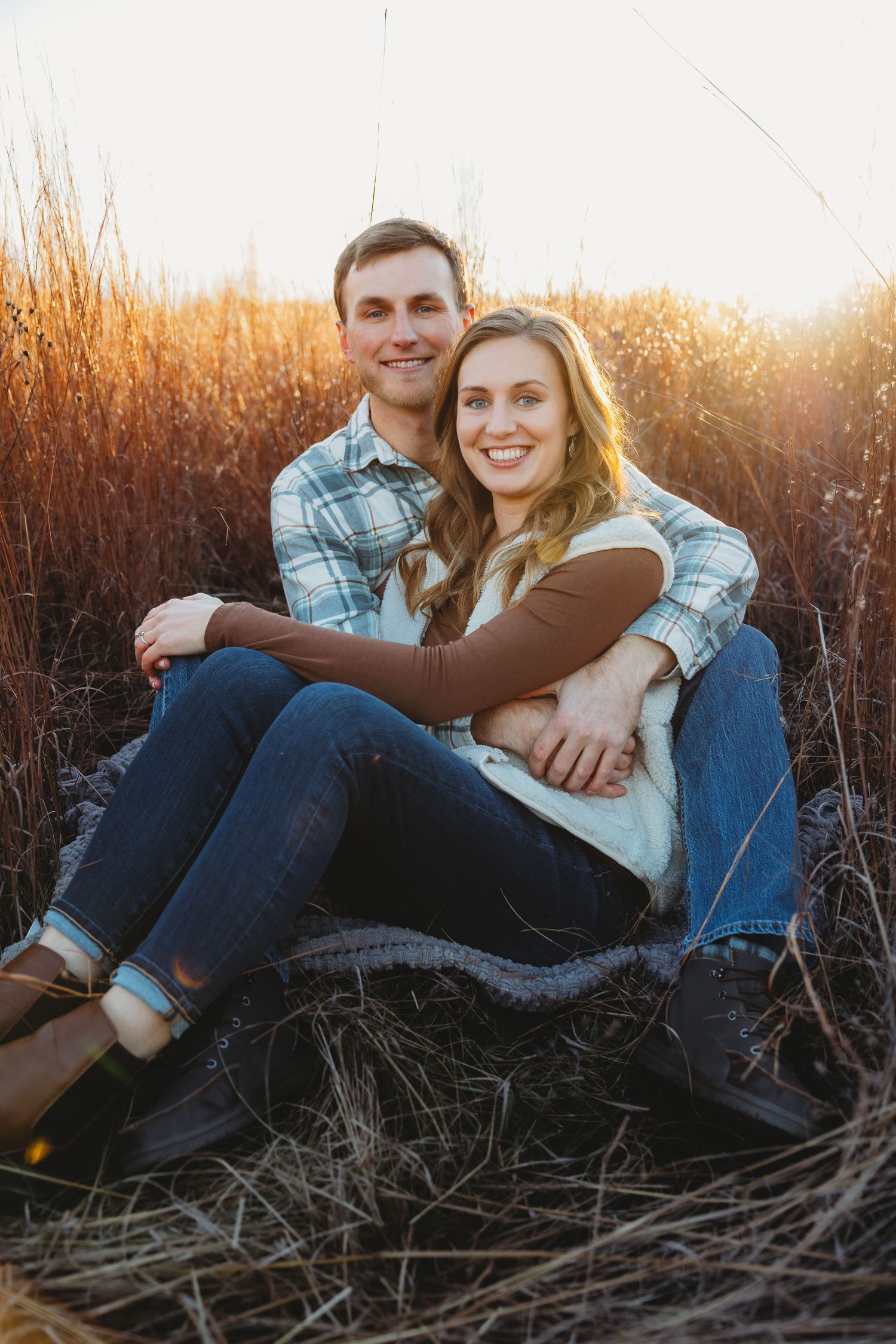 A couple relaxes together in tall grass during golden hour sunset, wearing casual denim and plaid clothing.